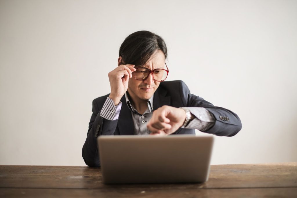 Asian businessman checking watch while working on laptop, expressing stress and urgency.